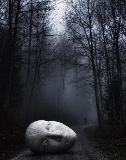 Photography and art. A color photo of a sculpture lying on a dark forest path. The sculpture is a large silver-colored head lying on its side. It is just a face, without hair, like a mask and looks a bit sad. The photo is very dark and some mist is rising between the trees. In the distance, a person dressed in dark clothes can be seen walking along the path.
Info: The 500-pound sculpture was made from packing paper and adhesive tape that the artist had left over when she moved to Los Angeles. It was used to protect the family property and had become superfluous. The "head" therefore stands for a new beginning. There was also a head lying next to fire during the photo session. Due to the events in Los Angeles with the terrible fire disaster in 2025, it suddenly took on a different meaning.