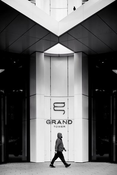 a darkly dressed man with a hood and a striking gait is photographed in the middle in front of the symmetrical entrance area of a high-rise apartment building with a white facade. the photo is very clean and symmetrical overall. this tidy look is cheekily ignored and loosened up by a dark dove sitting on the front roof.
