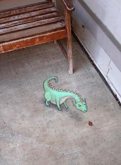 Streetart. A green dragon (named Alvin) and a red ladybug (named Celeste) have been drawn in chalk on a gray sidewalk next to a wooden bench. Big Alvin and the little Celeste are facing each other and staring at each other.
Title: "Alvin and Celeste's standoff has now entered its twelfth day, and neither can remember what the argument was about."