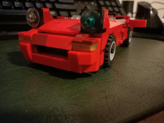 A LEGO model of a first-generation Mazda MX-5 NA, with the headlights up, sitting on a desk. The front and part of the left side are visible. The shot is taken from slightly above the car. There is no windshield on the MX-5, only a small 'stub' of transparent 1 by 1 wedge pieces, due to the limited amount of transparent parts available to me, and difficulty of creating such a windshield using other methods.