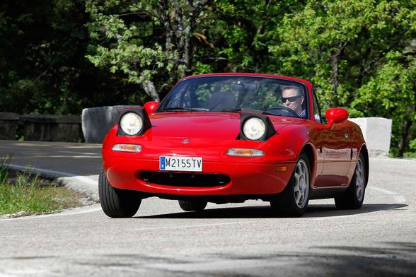 A picture of a first-generation Mazda MX5 NA provided for comparison with the pop up headlights up. It is pictured driving around a corner. The front and part of the left side is visible. It is shown at a similar angle to the first photo.