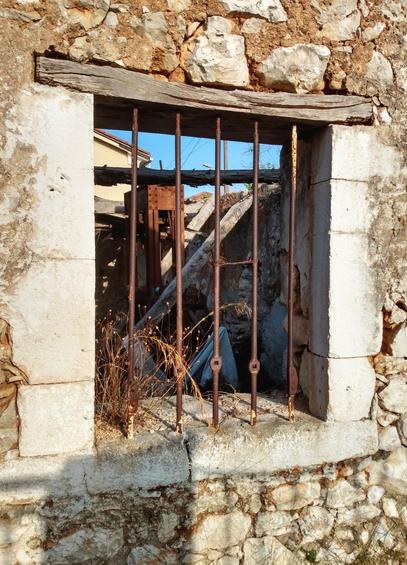 An old window in an abandoned old house. The frame is made of solid stone, only the beam above is made of weathered wood and surrounded by irregularly hewn fieldstones. The window has no glass or shutters, but five vertical rusty-brown, simply decorated metal struts run from top to bottom. The front of the house is lit by the sun. The interior of the house behind the window is untidy and loosely filled with junk. There is no longer a roof, so you can see a corner of the blue sky through the window.

Ein altes Fenster in einem verlassenen alten Haus. Der Rahmen ist aus massiven Steinen gemauert, nur der Balken oberhalb ist aus verwitterten Holz und umgeben von unregelmäßig gehauenen Feldsteinen. Das Fenster hat kein Glas und keine Fensterläden, aber von oben nach unten verlaufen fünf senkrechte rostig-braune, einfach verzierte Metallstreben. Die Hausfront ist von der Sonne beschienen. Der Innenraum des Hauses hinter dem Fenster ist unaufgeräumt und löse mit Gerümpel gefüllt. Ein Dach gibt es nicht mehr, sodass man durch das Fenster hindurch eine Ecke des blauen Himmels sehen kann.