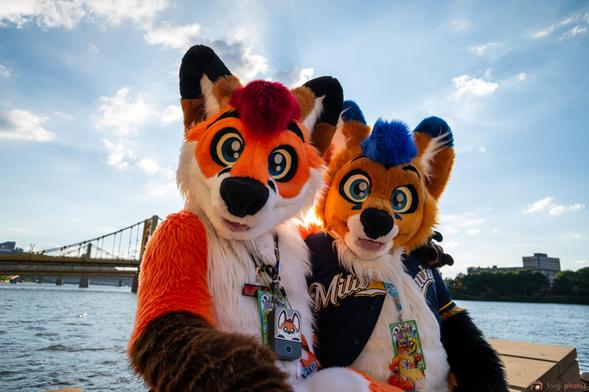 Colto Fox (orange, white, brown and red fox fursuiter) in full fursuit sitting on a pier while hugging Aero Fox (orange, white, black and brown fox fursuiter) also in full fursuit.

Behind them is a popular bridge in Pittsburgh, PA. They are down by the water in front of the convention centre at AnthroCon 2024.