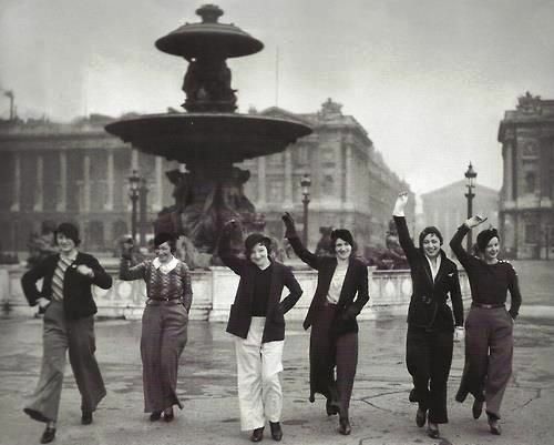 Six white women walking through Place de la Concorde in the late 1920s or early 1930s. They are all wearing trousers and laughing.