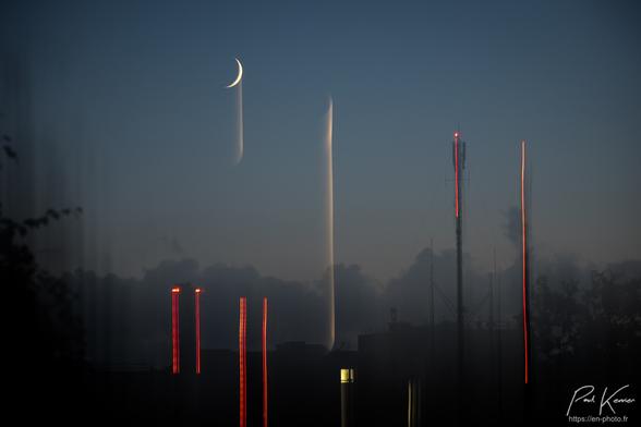 Photo en couleur, de nuit, d'une double captation en panotant verticalement. La silhouette du paysage, des arbres et le haut d'un bâtiment sont dans la pénombre, alors que le quartier de lune , les lumières de signalisation d'une antenne radio installée sur le toit du bâtiment d'une part et au sommet d'une cheminée d'autre part, ainsi qu'une fenêtre éclairée génèrent des filés colorés (blanchâtres pour la lune et la fenêtre et rouges pour les lumières de signalisation) et nettement plus clairs. Une des captations a été relativement brève en mouvement, et s'est terminée par un certain temps de stabilité, permettant de bien distinguer le quartier de lune et l'antenne sur fond de ciel dégagé. L'autre captation, décalée par rapport à la première prise, a été en mouvement tout au long de l'ouverture de l'obturateur et génère des filés fantomatiques nettement plus longs que ceux de la première captation.