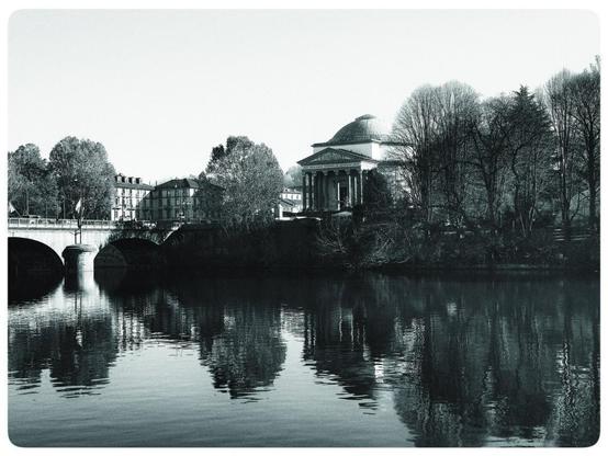 The image shows a black and white view of a neoclassical building with a dome, likely a church or a temple, by a river. There's a bridge in the foreground, connecting to the side of the river where the building is located. The trees along the riverbank are reflected in the water, and the overall setting appears serene and architectural.