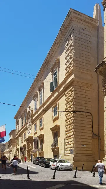 The image appears to depict a narrow street in a city with a notable building featuring a textured façade. The building seems to have multiple stories, and there are people walking along the street. The sky is clear and blue, suggesting it may be a sunny day. Additionally, there are decorative elements on the building, such as possibly sculptures or reliefs.

Image Credits: Wikimedia / Matthew Axiak / CC BY-SA 4.0