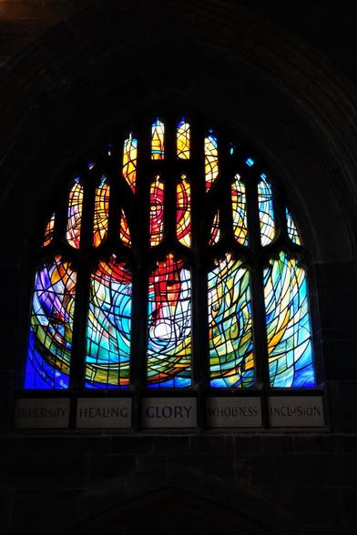 A large, colourful stained glass window with an abstract design and the words "Diversity," "Healing," "Glory," "Wholeness," and "Inclusion" written below.