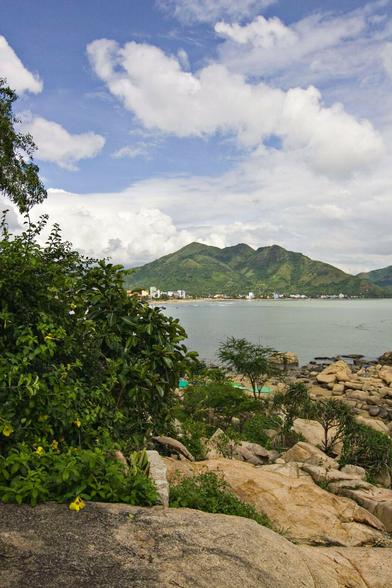 It's a bright, sunny, hot day with white puffy clouds in the blue sky suggesting high humidity. We're on the shore of what might be the sea or a lake with land far across the flat, grey-green water. Where we are there are large, sandstone boulders leading in a rough path down the water's edge with a few verdant shrubs poking up between gaps. A large bush grows on the left side forming a natural barrier there, and yellow flowers shine brightly in its dense, dark green foliage. The leaves of a tree just emerge top left from something just out of sight. The land over the water is jagged peaks, almost looking like a volcano carpeted in soft, green moss from this distance. Small blocks of whites and other pale colours line that distant shoreline; apartments and offices.