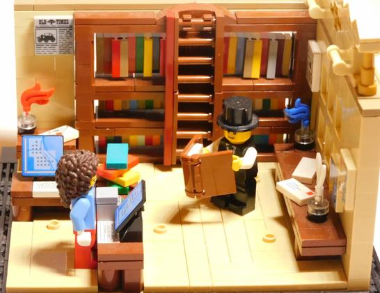 Lego model of a corner of a library showing a librarian at a counter with computers assisting a lego visitor in a top hat reading a book. The corers of the room are covered in a full book case
