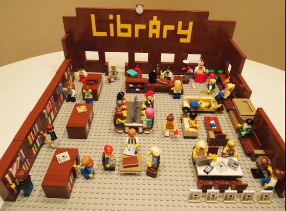 Lego display of a full library. I shows the word library in bricks at one end.  a staffed help desk. Patrons are sitting at several tables or on sofas reading. In one corner lego children are being read to by a lego chicken. There are several free standing book cases as well as book shelves along the left wall.