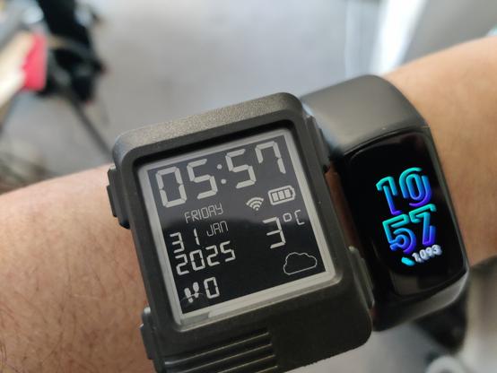 A Watchy watch showing the default screen, being worn on a wrist.  There is a Fitbit next to it. For scale, or just because I was too lazy to take it off? You decide!