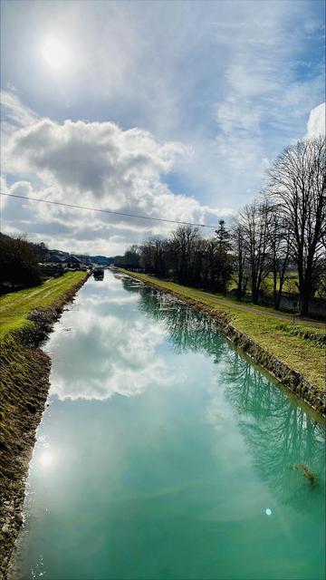 View onto the Canal de Bourgogne from a bridge. The water is turquoise and mirrors the very three-dimensional clouds and the winter sun breaking through with a bit of blue sky. Bare trees line the right shore, far away houses the left. A barge is parked further back.