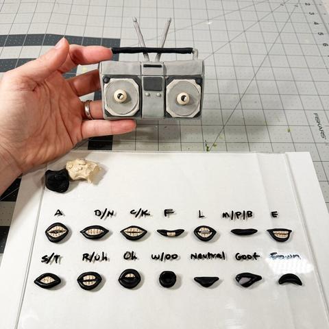 A hand holds a grey claymation boombox character. In the foreground, a set of mouths (aka visemes) are labeled on a piece of plexiglass.