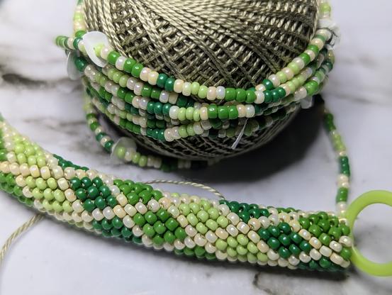 A beaded crochet rope in front of a small spool of attached crochet thread that is wrapped in a length of beads that have been prestrung for the pattern. The beads are three shades of opaque green along with an iridescent cream, and the pattern is rows of diamonds.
