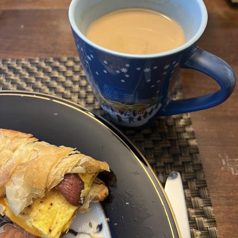 A close-up of a croissant sandwich filled with egg and bacon, served on a blue accented china plate. Next to it, a blue mug with a winter scene filled with creamed coffee.