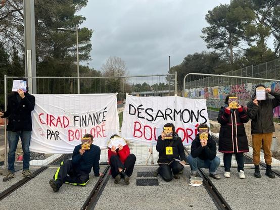 7 activistes au CIRAD, le visage caché par des tracts, devant des barrières où sont accrochés des banderoles "CIRAD financé par Bolloré" et "Désarmons Bolloré".