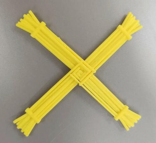 A yellow Brigid's Cross on a gray table. It's been painted a bit. Brigid's crosses are most often seen as braided wheat so the design attempts to mimic the look.