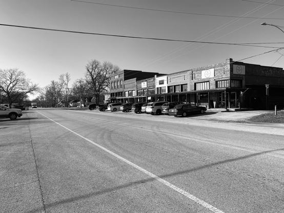 The images in black-and-white a powerline goes across the photo from side to side. In the foreground force perspective makes the highway look very wide. A series of mainly one story buildings in the style of the earliest 20th century is on the right side of the street. A line of carsis parked in front of them.