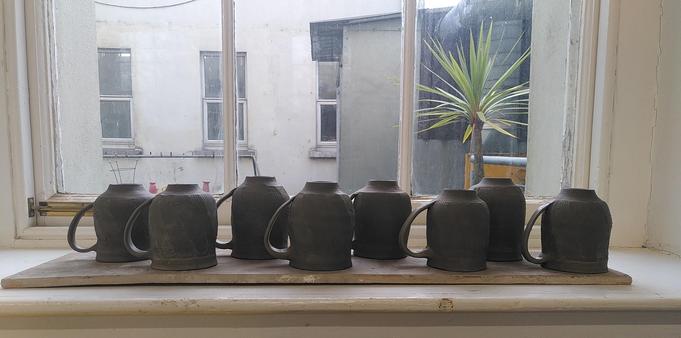 A line of ceramic mugs greenware stage upside down on a board to dry the newly made handles.
Une ligne de mugs en céramique encore frais étalés a l'envers pour faire sécher les anses tout juste posées.