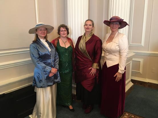 four women stand posed; left to right: Blue satin jacket with pearl buttons and a white satin full length skirt; Emerald green beaded silk two peice top and skirt; burgundy duopioni silk long jacket with gold silk collar and cuffs; off white silk jacket with pointed collar and cuffs over a dark plum velvet skirt.
the woman on each end waers a wide brimmed hat to match their outfits