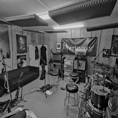 Das Schwarz-Weiß-Foto zeigt einen Proberaum für eine Band.  Der Raum ist relativ klein und mit schalldämpfenden Platten an der Decke und den Wänden ausgestattet.  Im Raum stehen mehrere Gitarrenverstärker, ein Schlagzeug, ein Sofa und diverse Musikinstrumente. An der Wand hängt ein Banner mit dem Bandnamen.  Poster mit Metal-Motiven schmücken die Wände.  Der Raum wirkt eher spartanisch eingerichtet, aber funktional.

Bereitgestellt von @altbot, generiert mit Gemini