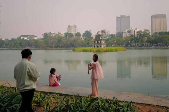 A woman in a pink dress and holding flowers, sitting close to the lake, being photographed by others.