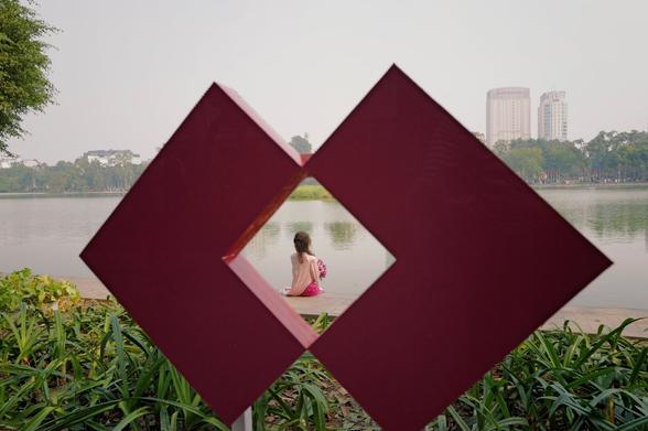 A woman in a pink dress sitting close to the lake. Around her as a kidn of frame is a big sign (?) in the foreground, that's also pink.