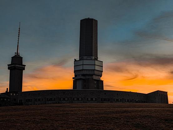 Großer Feldberg mit Sonnenuntergangshimmel: 
Das Bild zeigt die beeindruckende Silhouette des Großen Feldbergs, inklusive des markanten Fernsehturms und des Nebengebäudes, vor einem dramatischen Sonnenuntergangshimmel.
Der Himmel präsentiert sich in warmen Orangetönen, die sanft in dunkles Blau ubergehen. Die dunklen Konturen der Gebäude erzeugen eine kontrastreiche Stimmung und betonen die besondere Atmosphäre dieses Moments.