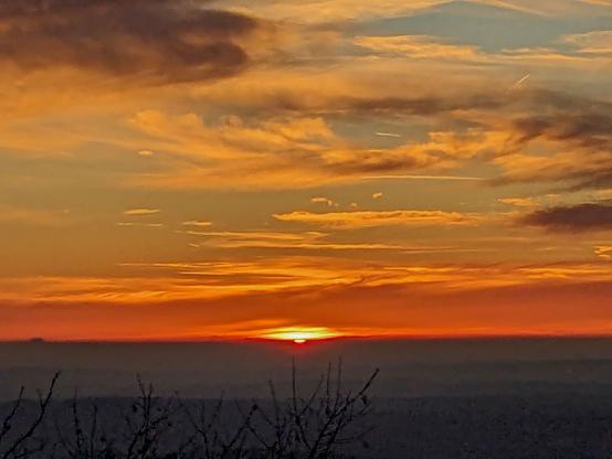Sonnenuntergang
über der Landschaft:
Hier steht der Sonnenuntergang selbst im Mittelpunkt. Die Sonne ist als roter Punkt am Horizont zu sehen, während der Himmel von intensiven Farben durchzogen wird - von tiefem Orange bis hin zu goldenen und blauen Nuancen.
Die dramatischen Wolkenstrukturen verleihen dem Bild Tiefe und verstärken die malerische Wirkung der Szene.