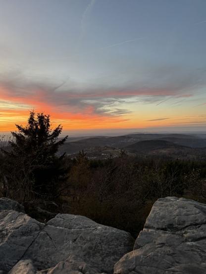 Blick vom Großen Feldberg ins Tal bei Sonnenuntergang:
Diese Aufnahme zeigt eine grandiose Panoramaansicht vom Feldberg ins Tal.
Im Vordergrund liegen felsige Strukturen, die einen natürlichen Rahmen für das Bild bilden. Links steht eine Tanne, die mit ihrer dunklen Silhouette den Blick weiter in die Weite der Landschaft lenkt.
Das Tal breitet sich unter einem Himmel aus, der von zarten Wolken und feinen Farbübergängen dominiert wird. Die warmen Farbtöne des Sonnenuntergangs reflektieren sich in der Landschaft und sorgen für eine harmonische Stimmung.