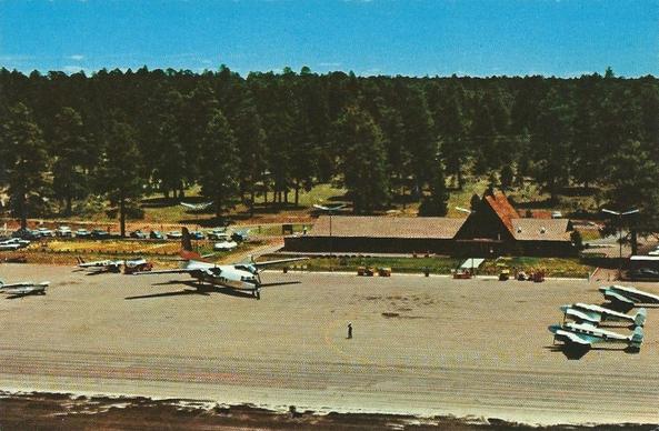 Overview of the airport reveals a simple airport building made out of wood. In the center is an A-frame. There is one large prop get to the left, and small ones on the right. You dont see the canyon , but you do see lots of trees.