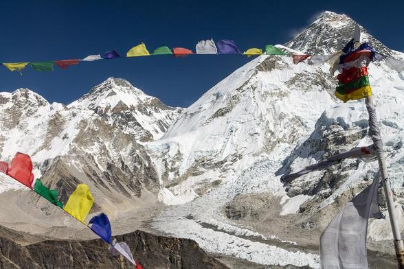 A colour photograph. There are colourful Buddhist prayer flags in the foreground and some snow-capped mountains including Mount Everest. – The classic view of Mount Everest (Chomolungma / Sagarmatha) from the top of Kala Patthar (5645m). The Khumbu Glacier with it famous Icefall is seen below and its bend is the place where tents of the Base Camp are set during the spring season. It is Changtse on the left, above Lho La which marks the border with Tibet. Autumn trekking season. Solukhumbum, Everest Base Camp Trek, Everest Region, Nepal.