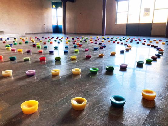 A big space with a natural stone tiled floor. Uniform, round blocks of candle wax in various colors (red, green, pink, purple, blue, yellow etc) are placed on the floor in concentric circles.