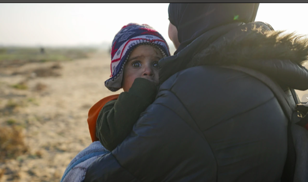 A imagem mostra uma criança de colo, com um gorro de tricô colorido e o que parece ser um agasalho escuro, levada nos braços de uma mulher adulta. A criança tem os olhos escuros muito abertos e fixos, olhando na direção da câmara, parecendo estar amedrontada, escondendo o rosto parcialmente com as mãos. A mulher usa um casaco escuro, acolchoado, com a cabeça coberta por lenço ou gorro igualmente escuro vendo-se as alças da mochila escura que leva às costas. Ambos caminham num terreno árido, aparentemente junto ao litoral, onde se vislumbra o que parecem ser ruínas ou edifícios demolidos. A fotografia foi tirada no Norte de Gaza, durante o cessar-fogo que permitiu o regresso de milhares de famílias palestinianas forçadas a evacuar as suas casas sob os bombardeamentos israelitas.