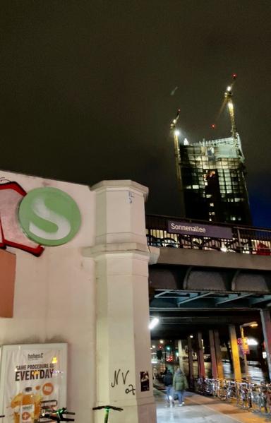 Das Nachtbild zeigt einen Teil einer Stadtlandschaft. Im Vordergrund steht eine weiß verputzte Säule, an der ein grün-weißes Kreislogo mit einem „S“ zu sehen ist, sowie Graffiti und ein Werbeschild für ein Getränk. Hinter der Säule verläuft eine Brücke über eine Straße, auf der sich Fahrräder befinden und Menschen zu Fuß unterwegs sind. Im Hintergrund ist ein Hochhaus im Bau zu sehen, an dem Baukräne angebracht sind. Am Ende der Brücke steht der Name „Sonnenallee“ auf einem Schild. Der Himmel ist dunkel und bewölkt.

Bereitgestellt von @altbot@fuzzies.wtf, generiert mit Gemini