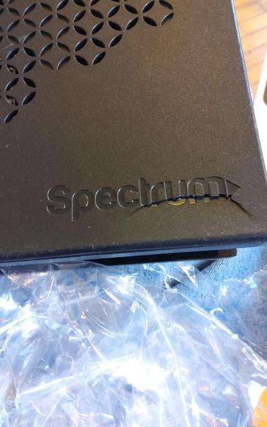 A freshly unwrapped Spectrum cable modem.  The shrinkwrap is at the bottom of the picture.  The outside of the modem is black plastic with air holes at top left.  At the bottom right, the cable company name, Spectrum, is stamped into the plastic.  A semi-circular crack extends from the bottom right corner of the modem, up into the name, and then curves back down to the left, back below the name.