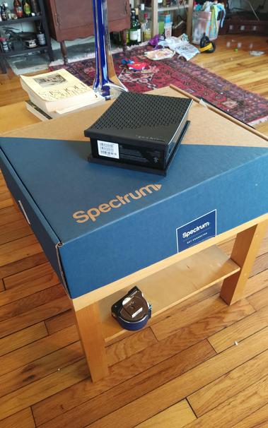 The giant box delivered by FedEx containing the new, yet physically broken cable modem (on top of the box).  The box is on my coffee table.  The box also contained some pieces of paper, 2 small cables shrink-wrapped together, and a power adapter.  Below the box on a shelf is the tape measure I used to measure the box before taking out all the stuff.  It measured about 2112 cubic inches.  I imagine the modem was broken before it was shrink-wrapped, but ¯⁠\⁠_⁠(⁠ツ⁠)⁠_⁠/⁠¯
