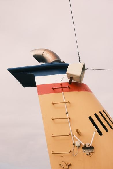 This image shows the smokestack or funnel of a ship. It has a distinct yellow, red, and blue color scheme. The funnel is equipped with an exhaust pipe and has an attached vent or speaker. There are also steps or ladder-like structures for access. The setup likely includes various utility components typically seen on a ship's funnel.