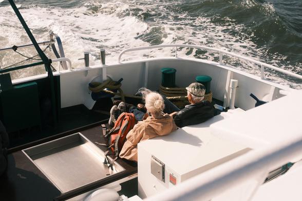 The image shows two people sitting on the deck of a boat, looking at the ocean waves. They appear to be relaxing, with some bags near them. The scene includes safety equipment and ropes on the deck. The water is choppy, suggesting the boat is moving.