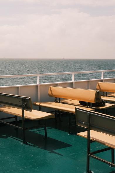 The image shows a view from a ferry or a boat's deck. There are empty benches arranged for passengers with the ocean and sky in the background. The deck has a green floor, and the benches are wooden with metal supports.