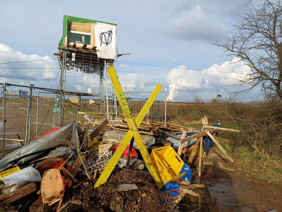 Eine Hütte auf einer Barrikade der Werkstraße am Rand des Tagebau Hambach beim Manheimer Erbwald. Im Vordergrund ein großes gelbes X aus Holz. Im Hintergrund qualmende Kühltürme im Rheinischen Revier.