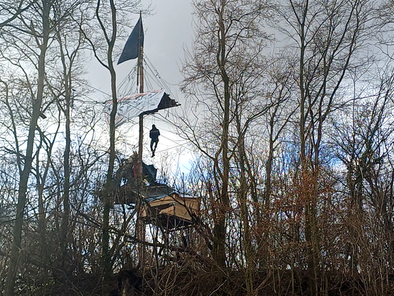 Waldbesetzer*innen klettern an Seilen auf einer Plattform, über der eine schwarze Fahne weht.
