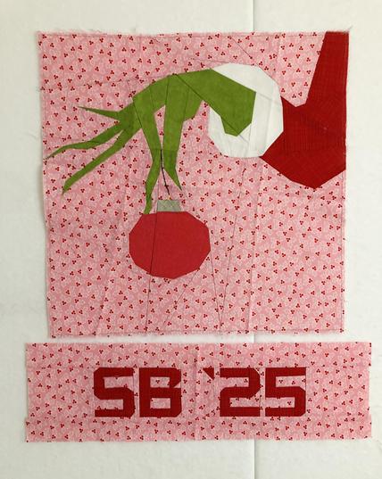 An intricately pieced FPP quilt block of the Grinch’s hand dangling an ornament as if he’s about to let it fall. The hand is Grinch green in a red and white Santa sleeve and the ornament is red, as well. The background is a pink holly ditsy fabric. A separate block has pieced letters, in red on the pink holly, that read “SB ‘25”