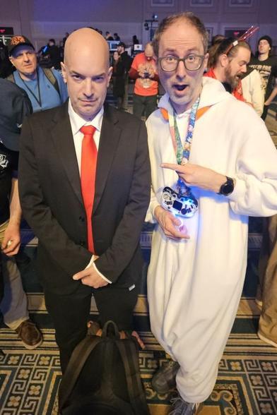 A serious looking bald man in a nice suit and red tie next to a less serious, less bald man wearing a Scorbunny kigurumi.