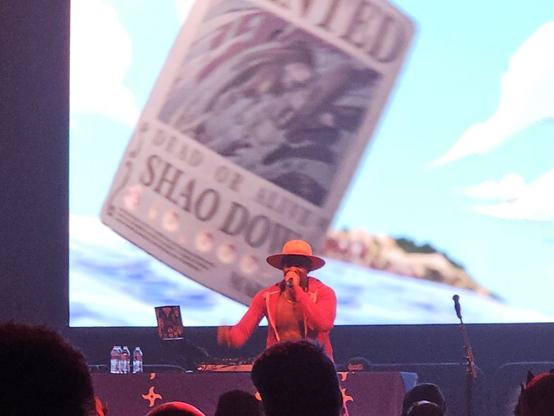 A man in a Monkey D. Luffy cosplay raps in front of a screen showing a Marine wanted poster with Shao Dow having a 10,000 Berry bounty.