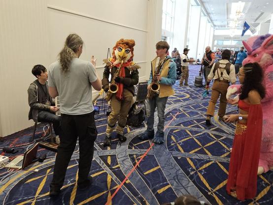 A crowded hallway. In the center, a person in bird fursuit & another person hold saxophones. Other musicians left, onlookers on right.