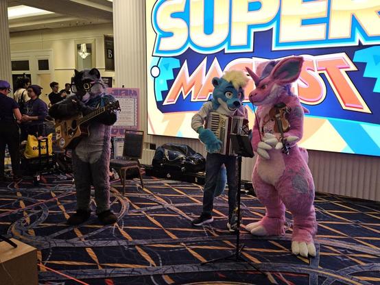 Three fursuiters -- raccoon, otter, and nidorino from Pokemon -- in front of a large Super MAGFest sign, holding musical instruments.