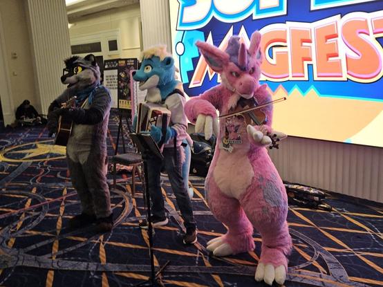Three fursuiters -- raccoon, otter, and nidorino from Pokemon -- in front of a large Super MAGFest sign, performing a song.