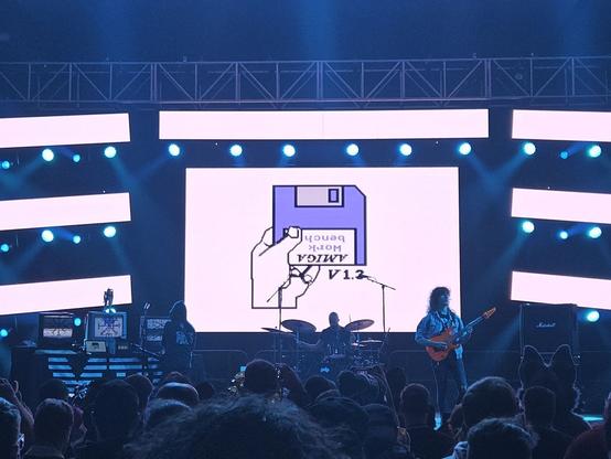A large screen behind a stage. On the screen is a hand holding an upside down floppy disk that reads "Amiga Workbench V1.3".